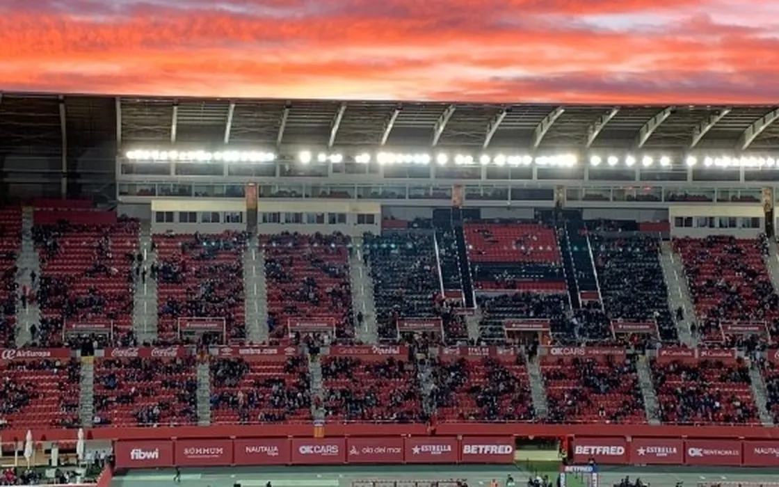 Tribüne des Stadions von RCD Mallorca bei Sonnenuntergang, mit rotem Himmel und eingeschaltetem Flutlicht.